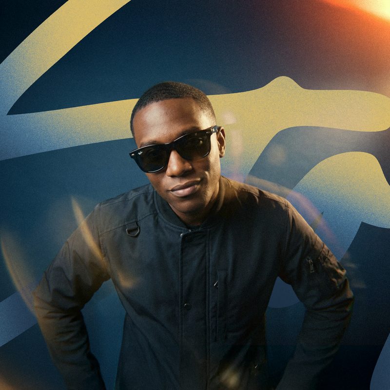 A cool Black man in a black bomber jacket and dark Wayfarer sunglasses looks directly at the viewer with a slight smirk. He stands against a backdrop of sweeping, abstract yellow and blue shapes, with warm lens flares emanating from the top right and bottom left, creating a vibrant, energetic mood.