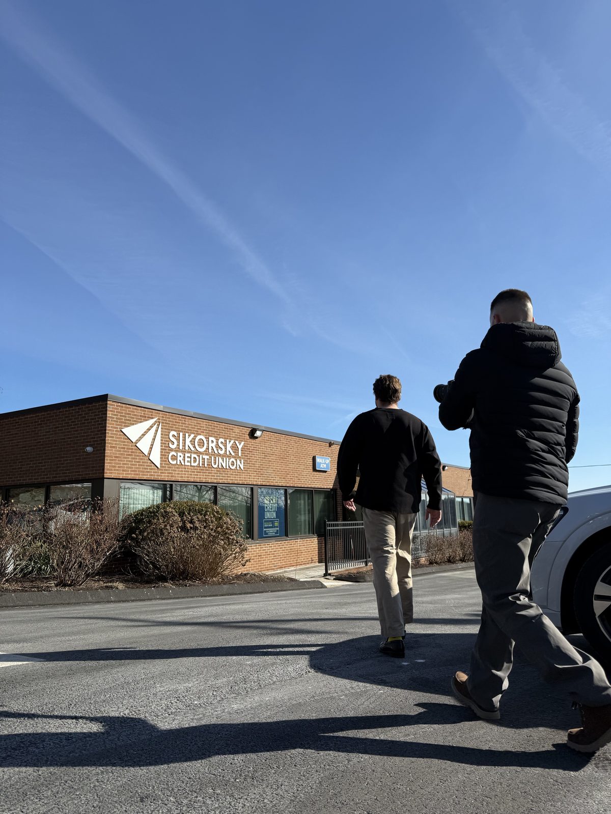 Two men walk across an asphalt lot towards the brick exterior of Sikorsky Credit Union on a bright, sunny day. The man in the foreground, wearing a dark puffer jacket, casts a long shadow as he approaches the building, which features large windows and a clear blue sky overhead. The mood is clear and purposeful, as if on a site visit.