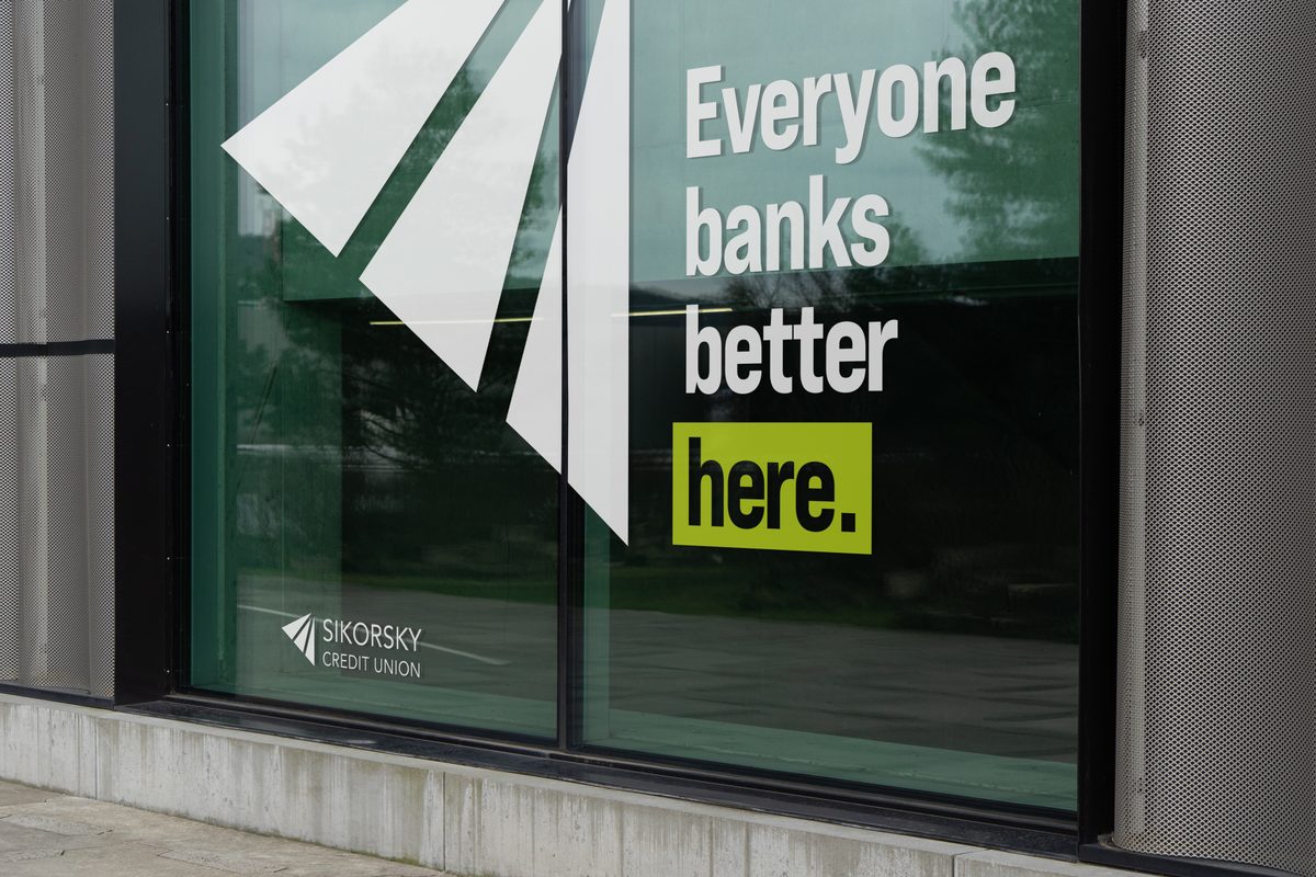 A modern bank window displays "Everyone banks better here." in white and a bright green box, with a white abstract design and the Sikorsky Credit Union logo. The outdoor scene is reflected in the window, which is set above a concrete base and flanked by a gray perforated metal wall.