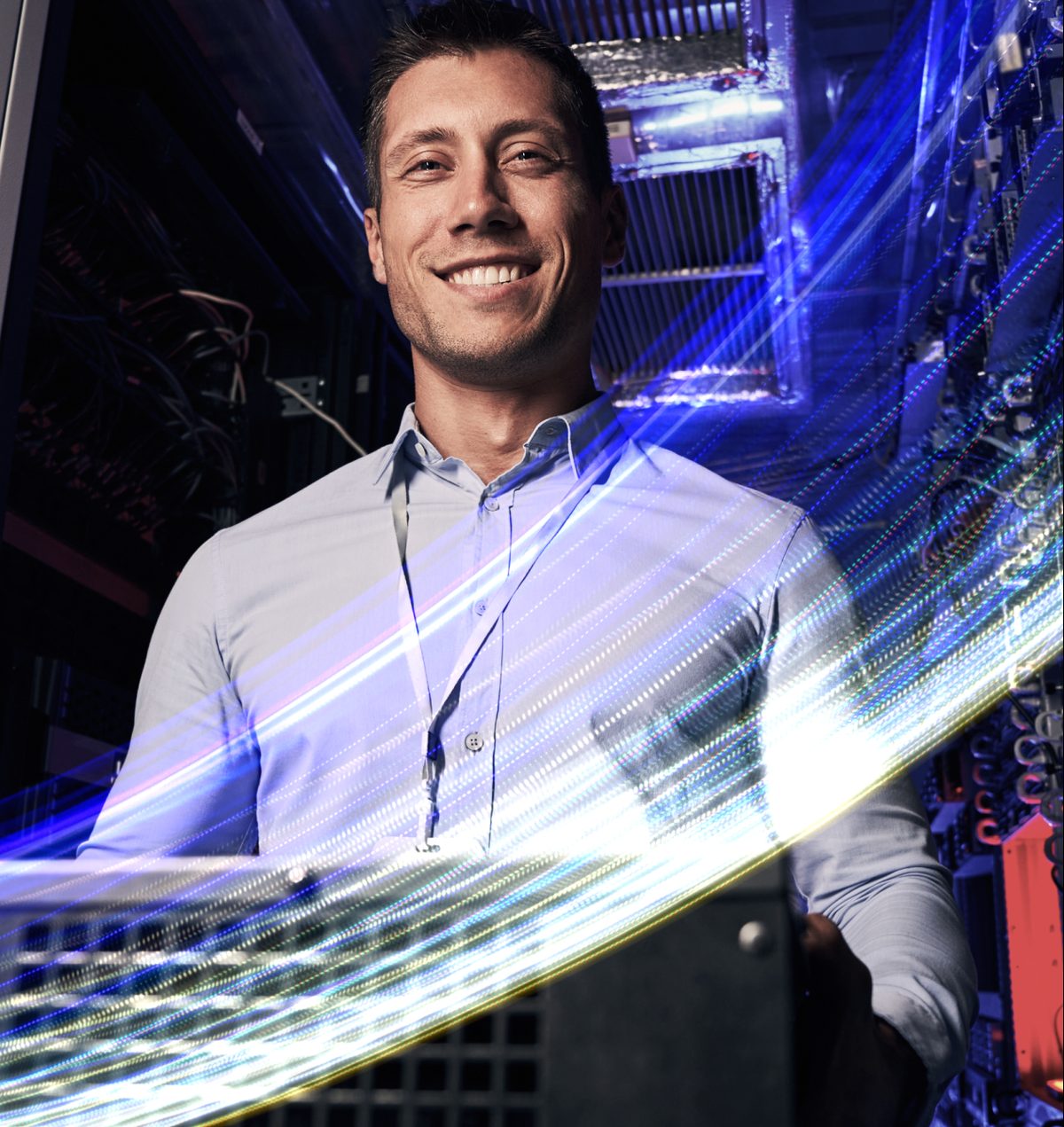 A smiling man in a light blue button-down shirt stands in a dim server room, illuminated by the bright blue and yellow light streaks of digital data flowing around him. He wears a lanyard and looks confidently at the viewer, conveying a mood of technological expertise and professionalism.