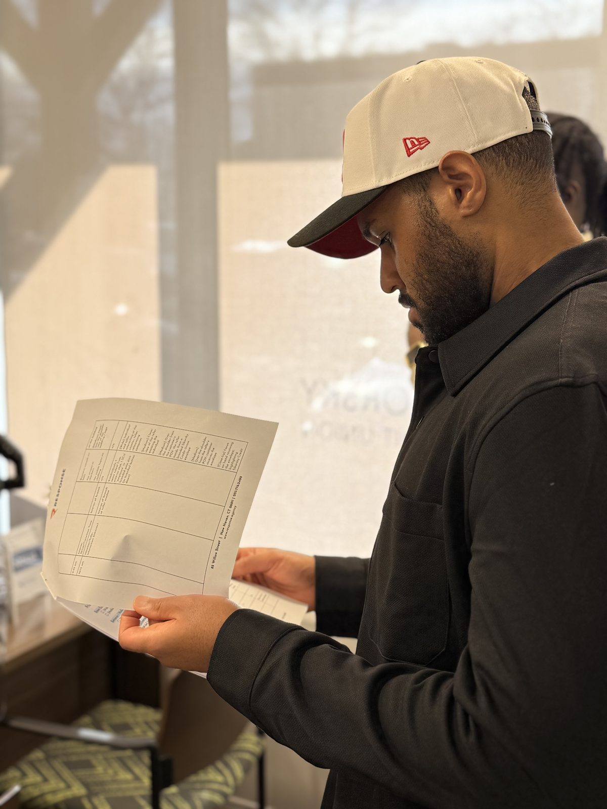A focused individual in a cream-colored baseball cap with a red brim and logo, and a black collared shirt, intently reads a white document. Their hands delicately hold the paper, highlighting the detailed text on the sheet. The background is softly blurred, with hints of a brightly lit interior, suggesting a moment of quiet concentration amidst an active environment.