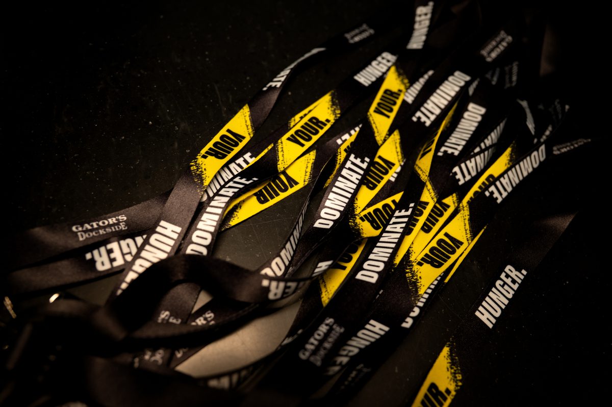 A pile of black lanyards with bold white and yellow text reading "DOMINATE. YOUR. HUNGER." sprawled across them, suggesting an energetic, competitive mood. Some lanyards also feature the "Gator's Dockside" logo in a smaller white font. The scene is dimly lit with a dark background, making the vibrant yellow and white pop.