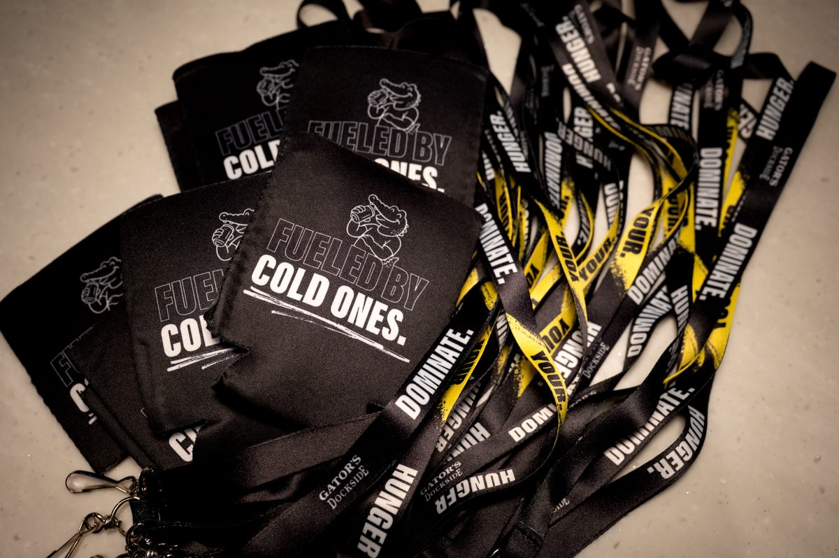 A close-up shot of several black koozies and lanyards. The koozies are printed with "FUELED BY COLD ONES." and a cartoon alligator mascot, while the lanyards feature "DOMINATE," "HUNGER," and yellow hazard-stripe accents. The items are arranged on a light-colored surface with warm, soft lighting, conveying an edgy and energetic brand aesthetic.