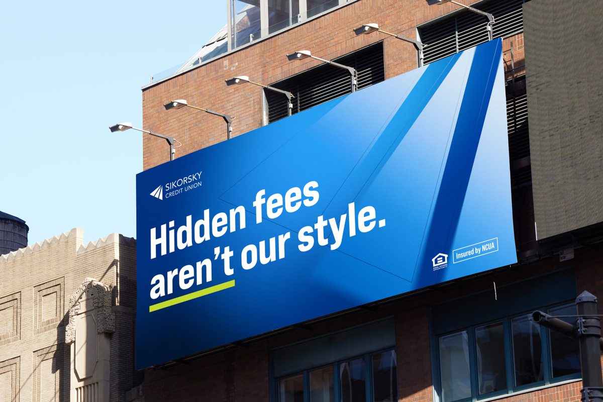 A large billboard for Sikorsky Credit Union is prominently displayed against a clear blue sky and brick buildings, featuring the slogan "Hidden fees aren't our style." The design uses various shades of blue with a modern, geometric aesthetic, and the text "Insured by NCUA" is visible in the bottom right corner. Bright daylight illuminates the scene, casting subtle shadows and highlighting the texture of the surrounding architecture.