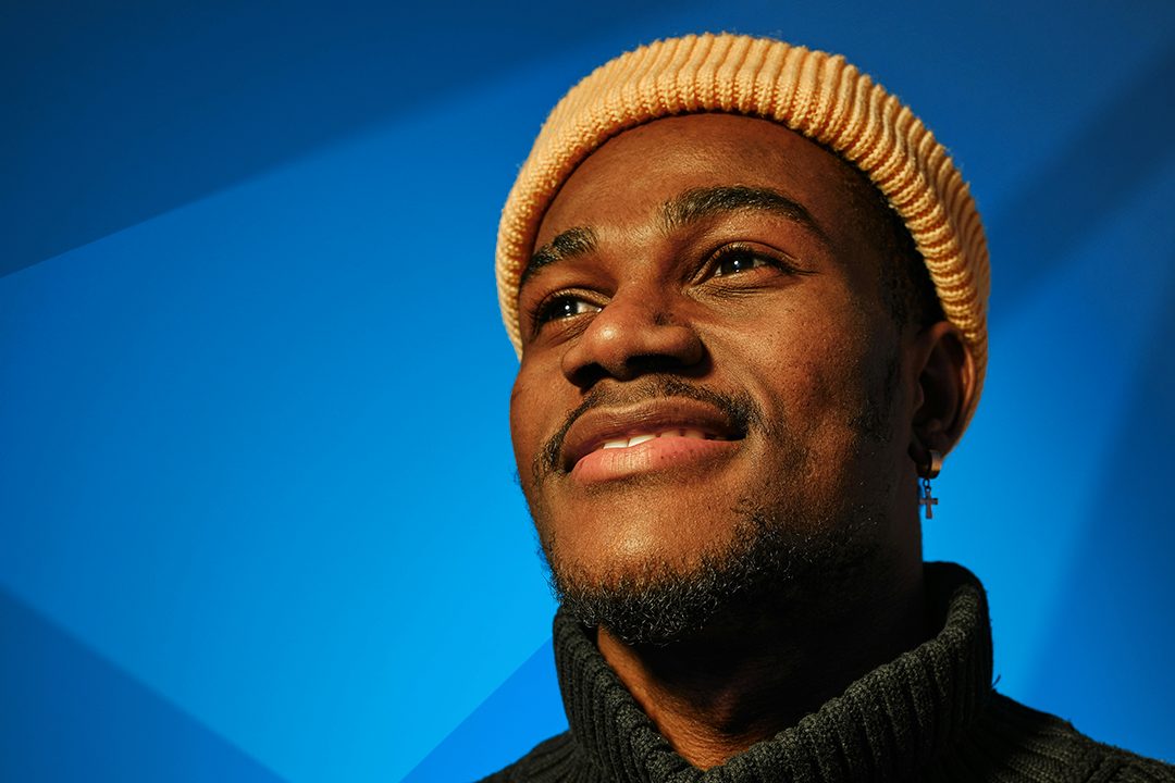 A smiling Black man, wearing a beige knit cap and black turtleneck, gazes upwards with a hopeful expression against a vibrant blue background. Dramatic lighting casts a sharp shadow diagonally across the upper left and right, highlighting his smooth skin and the subtle texture of his clothing.