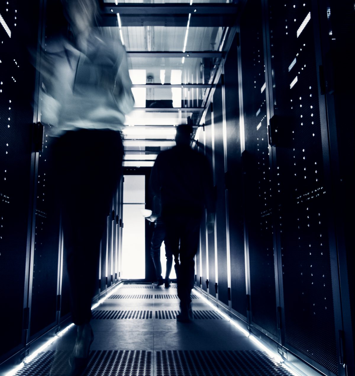 Four blurred figures move rapidly through a dark, high-tech server room, bathed in cool blue and white light. The motion blur on the two foreground figures creates a dynamic sense of urgency as they navigate the glowing data racks. The scene evokes a mood of intense activity and digital connectivity.