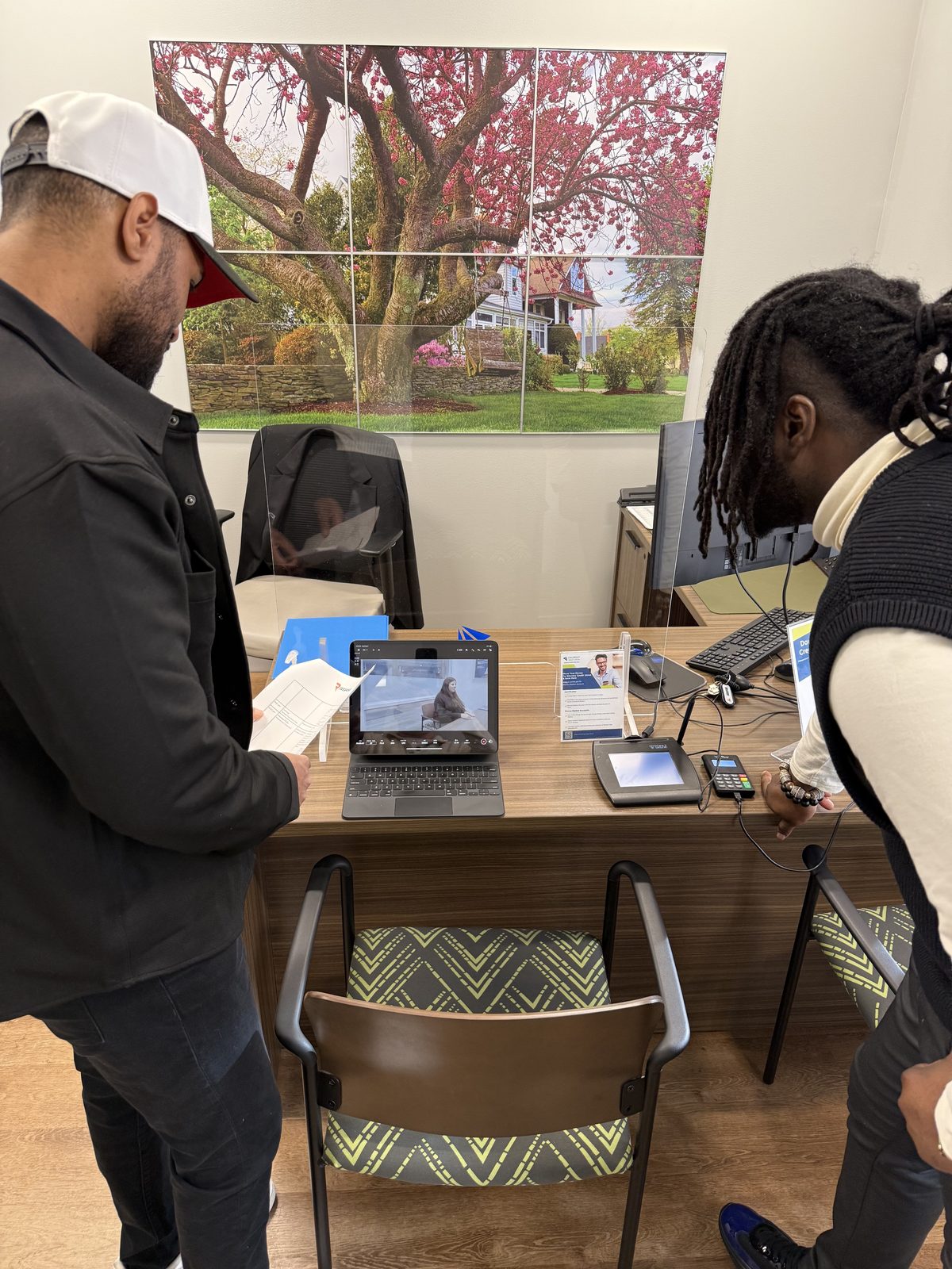Two men lean over a wooden desk, engrossed in a video call displayed on a laptop, with a large, vibrant mural of a cherry blossom tree and a quaint house behind them. The man on the left, wearing a white baseball cap and dark jacket, holds papers, while the man on the right, with dreadlocks and a light-colored sweater, reaches toward a payment terminal. The scene feels modern and collaborative under bright, even lighting.