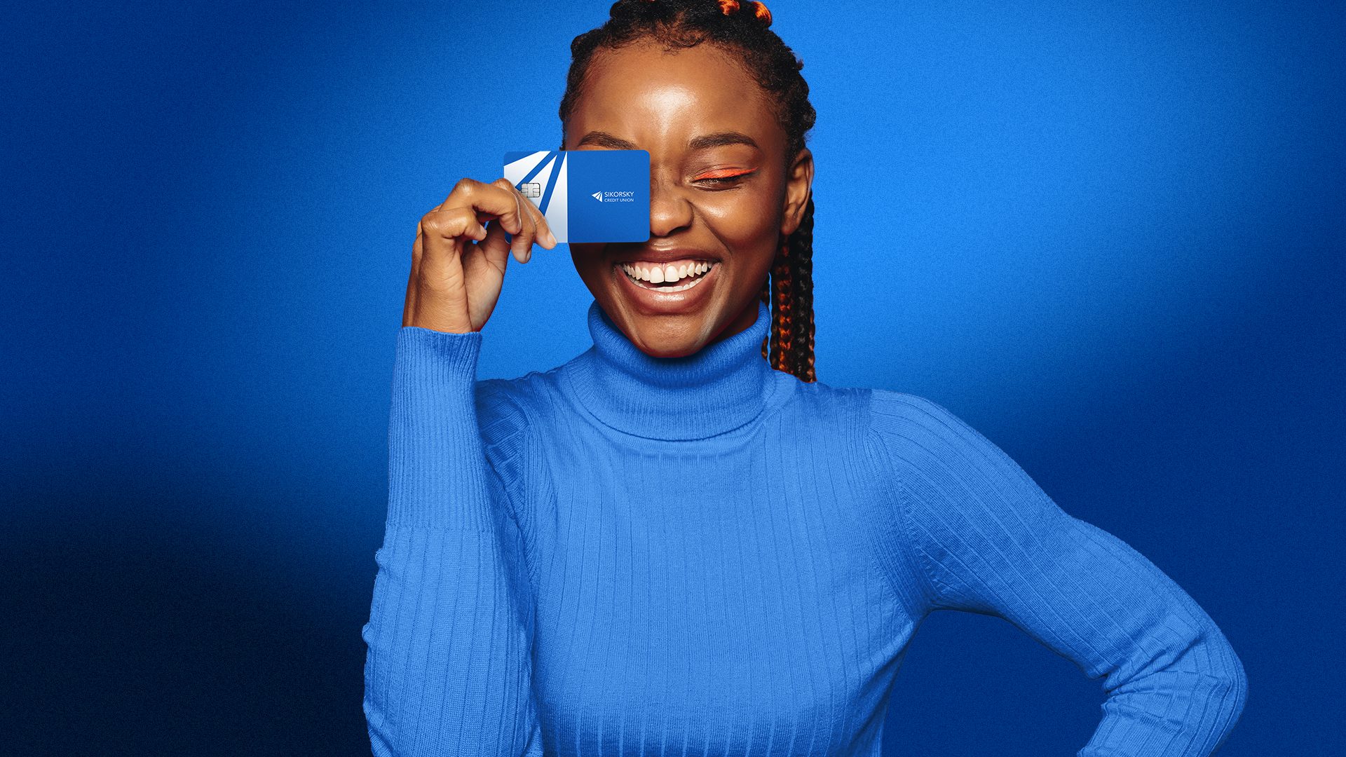 A joyful woman with dark braided hair and a royal blue ribbed turtleneck sweater smiles widely, covering one eye with a blue and white Sikorsky Credit Union card. Her eye makeup is a bright orange winged liner, contrasting against the vibrant blue background. The lighting highlights her face and the card, creating a playful, optimistic mood.