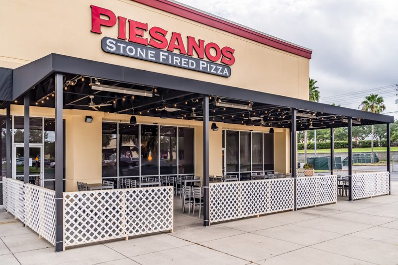 The front exterior of "Piesanos Stone Fired Pizza" restaurant is shown under a dark sky, featuring a large black awning strung with small lights over a spacious outdoor patio. The patio is enclosed by a white lattice fence and furnished with numerous tables and chairs, suggesting a welcoming, casual dining experience.