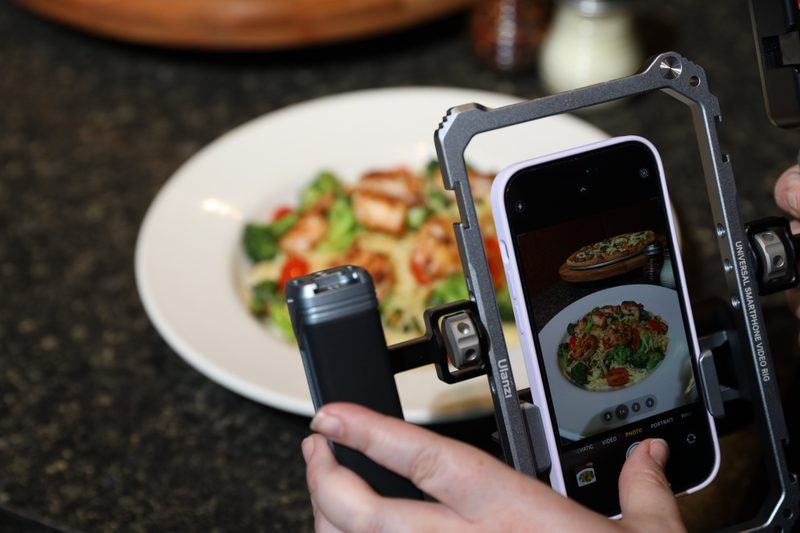 A person holds a phone in a "Universal Smartphone Video Rig" over a blurred plate of food. The phone displays the same plate of food in focus on its screen, along with another picture of a pizza. The lighting is bright on the food, indicating a well-lit food photography setup.
