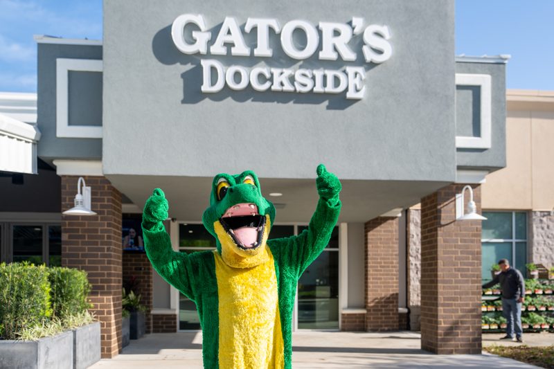 A cheerful green and yellow alligator mascot stands front and center, giving two thumbs up with a wide, open-mouthed smile. Behind it, the "GATOR'S DOCKSIDE" sign in large white letters casts a crisp shadow on a gray building, while warm sunlight bathes the scene.