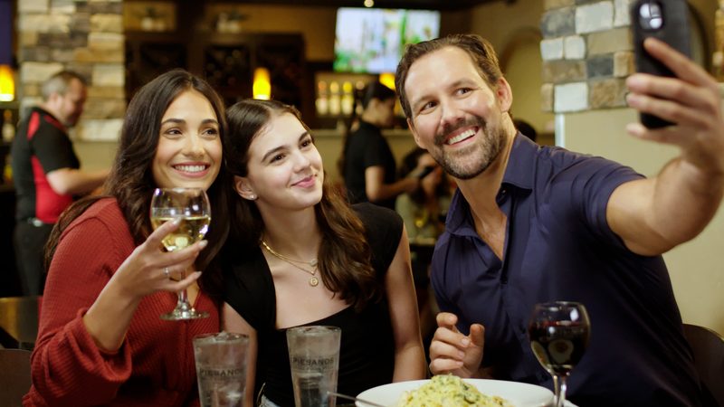 A family of three, a smiling man, woman, and teenage girl, takes a selfie at a restaurant table. The man holds a phone up with his right hand as the woman holds a glass of white wine and the teen smiles warmly, with a plate of pasta and two glasses of water visible on the table.
