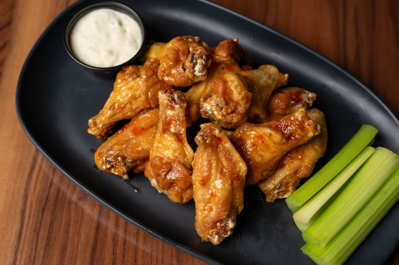 A dark, oval plate holds a pile of glistening, golden-brown chicken wings with visible bits of red spice and sauce. To the left is a small black cup of creamy white dipping sauce, and to the right are several bright green celery sticks. The plate rests on a warm, brown wooden surface.