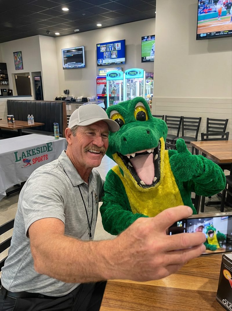 A man with a mustache and a grey baseball cap smiles while taking a selfie with a bright green and yellow alligator mascot, who gives a thumbs up. They are in a well-lit, modern restaurant with multiple TVs showing sports and claw machines in the background. The phone screen reflects the joyful selfie, capturing the fun, celebratory mood.