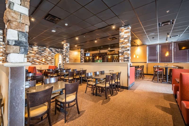 A warmly lit, inviting restaurant interior with a dark ceiling and an earthy color palette. Stone columns and accent walls with warm orange booths and tables with dark wood chairs are arranged on a light brown carpet. A bar area with hanging pendant lights and high-top tables can be seen in the background.