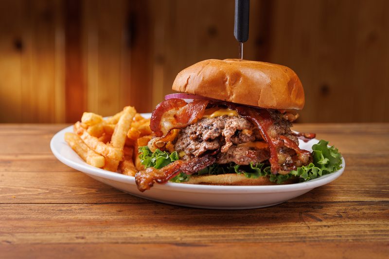 A towering bacon cheeseburger, speared with a black knife, sits on a white plate with a side of golden fries. The burger features melted cheese, crisp bacon strips, fresh lettuce, and red onion, all on a toasted bun, resting on a rustic wooden table with a blurred warm wooden wall in the background. The low, warm lighting creates an inviting and mouthwatering mood.